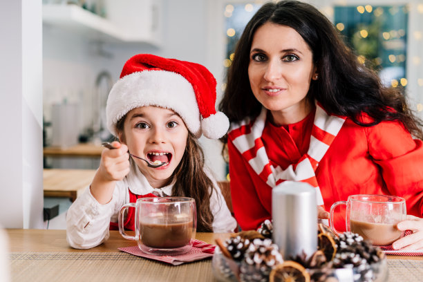 戴着圣诞帽的小女孩和她的妈妈坐在装饰精美的现代厨房里，喝着可可和棉花糖。家庭度假。喜庆的圣诞气氛。有选择性的重点。图片下载