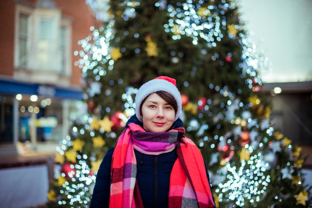 在空旷的街道上，戴着圣诞帽的混血妇女在装饰好的户外圣诞树的背景下。流行病期间的圣诞节概念。选择性聚焦，复制空间。图片下载