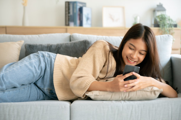 快乐的年轻亚洲女性放松在舒适的沙发上在智能手机上发短信，微笑的女孩使用手机，浏览无线互联网上的小工具，从家里网上购物图片下载