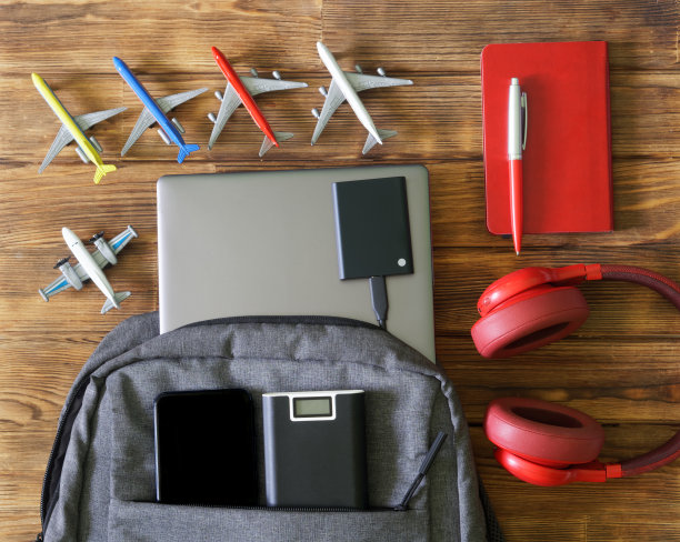 灰色背包，木制背景的笔记本电脑，ssd硬盘，手机和充电宝，旁边是耳机，记事本，钢笔和玩具客机。图片下载