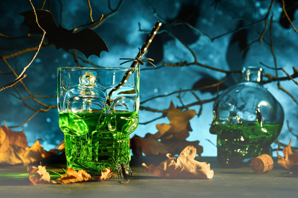 万圣节血腥的鸡尾酒。万圣节绿色鸡尾酒玻璃高脚杯和瓶子形状的头骨在黑暗的背景。有糖果眼睛，蝙蝠，蜘蛛的万圣节派对。亡灵节。Clous-up。图片下载