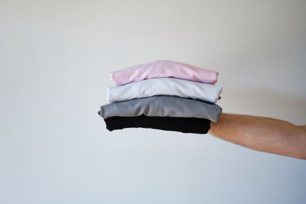 一叠干净的、刚洗过的、叠得整整齐齐的衣服在明亮的背景下拿在手里。白色墙壁背景上的一堆五颜六色的t恤。复制空格，关闭图片下载