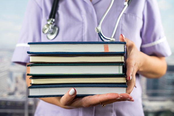 裁剪图像的实习生在医疗制服拿着许多书在她在医院的前面。图片下载