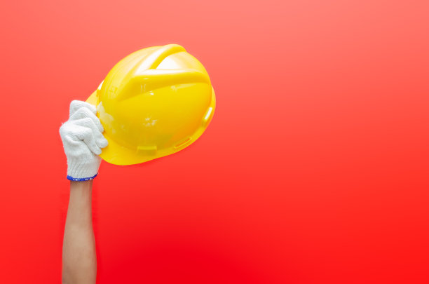 男性手戴手套，手持黄色安全帽，红色背景隔离。安全生产的概念。劳动节的概念。图片下载