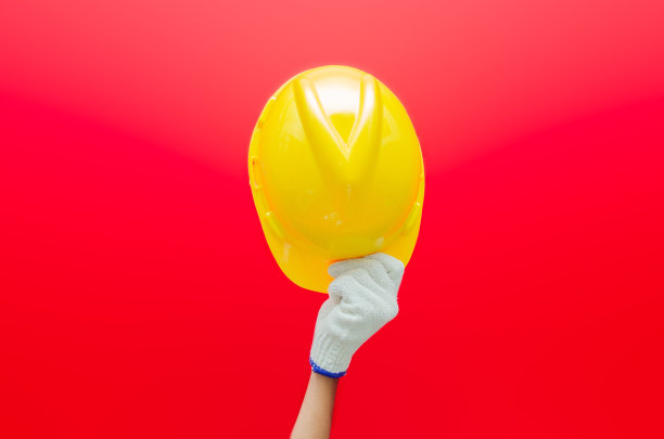 男性手戴手套，手持黄色安全帽，红色背景隔离。安全生产的概念。劳动节的概念。图片下载