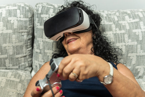 一名戴着虚拟现实眼镜的卷头发黑发老年妇女正在玩游戏，一边转着头一边转动着游戏机手柄。图片下载