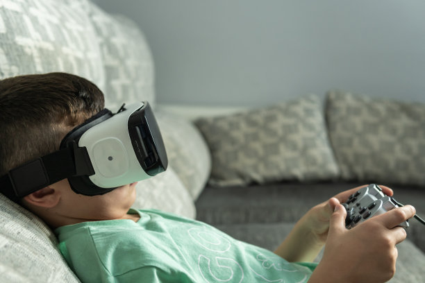 一个带着虚拟现实眼镜的男孩斜靠在沙发上用控制器玩在线游戏的侧面图像。图片下载