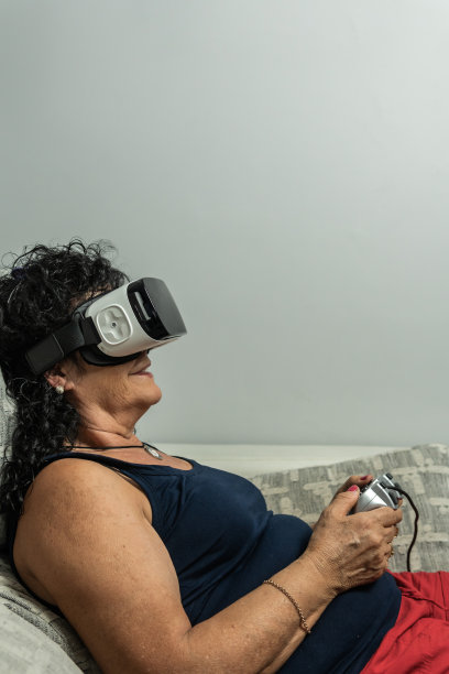 垂直拍摄的卷发黑发高级妇女戴着虚拟现实眼镜和手持游戏机控制器，而躺在沙发上。图片下载