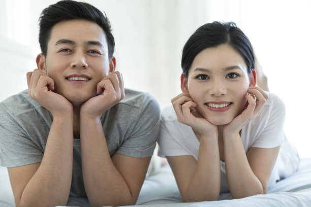 正面肖像的年轻夫妇躺在卧室的床上，手拿着下巴像两朵花-库存照片图片下载