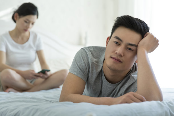 年轻夫妇或坐或卧在床前，妻子低头，丈夫表情阴沉。情感上的脱节——库存照片图片下载