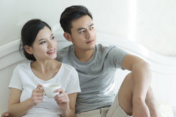 年轻的夫妇坐在卧室的床上望着窗外，妻子端着热饮——库存照片图片下载