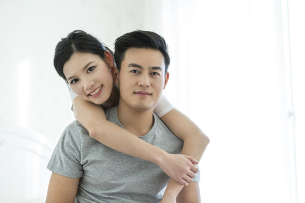 年轻的东亚华人夫妇的肖像在卧室床上穿着家居服，妻子与她的手臂环绕她的丈夫从后面-库存照片图片下载