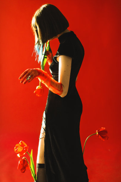 深色头发的亚洲妇女与郁金香在手与烟在工作室红色背景。时尚模特，白皙的皮肤，棕色的头发，黑色的衣服。手套的手。风格图片下载