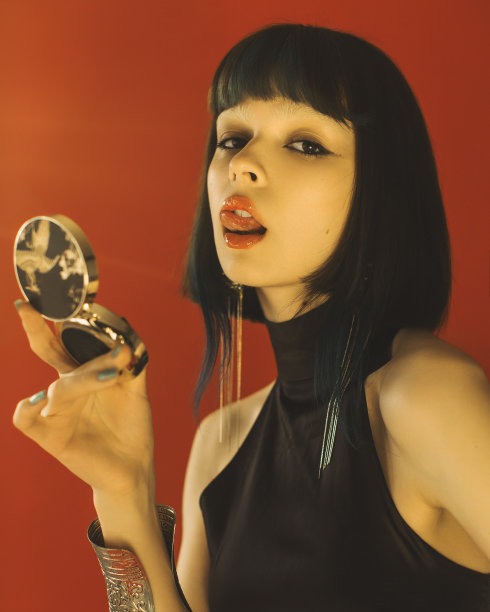 美丽的肖像。发型。褐发亚洲妇女与镜子在手在工作室的红色背景摆姿势。时尚模特，白皙的皮肤，棕色的头发，黑色的衣服。风格。耳环图片下载