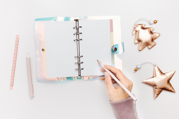 新年计划概念，女人拿着铅笔，写在打开空白的抄写本或日记本上。粉色金属色圣诞树和闪亮的星星玩具。学校或家庭办公文具图片下载