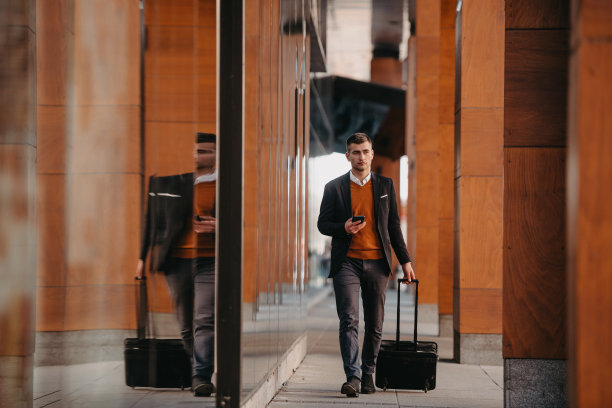 去机场候机楼。自信的商人旅行者走在城市的街道上，拉着他的行李箱，喝着咖啡，用智能手机说话图片下载