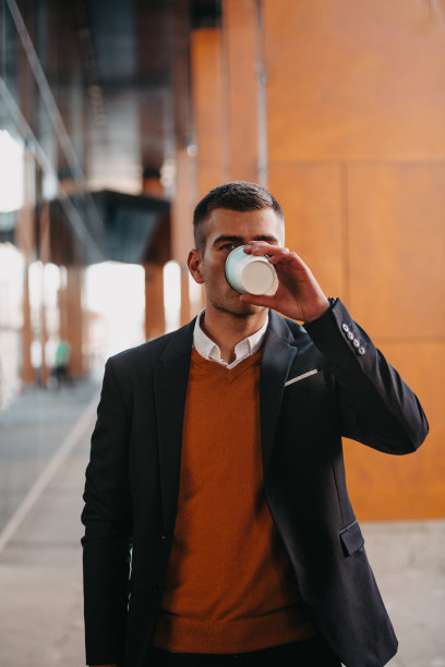 去机场候机楼。自信的商人旅行者走在城市的街道上，拉着他的行李箱，喝着咖啡，用智能手机说话图片下载