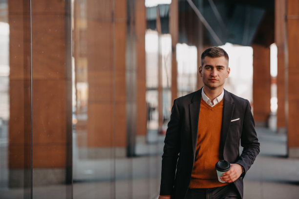 去机场候机楼。自信的商人旅行者走在城市的街道上，拉着他的行李箱，喝着咖啡，用智能手机说话图片下载