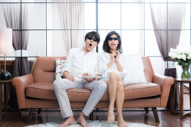 年轻的东亚夫妇在客厅沙发上看3D电影-库存照片图片下载