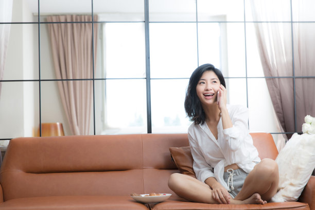 年轻的东亚妇女坐在客厅沙发上用她的智能手机打电话-库存照片图片下载