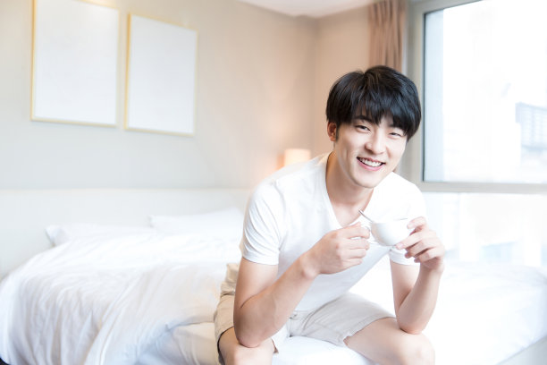 年轻的东亚人坐在现代家庭卧室的床上喝咖啡-库存照片图片下载