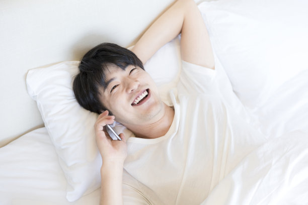 年轻的东亚男子使用智能手机躺在现代家庭卧室的床-库存照片素材图片