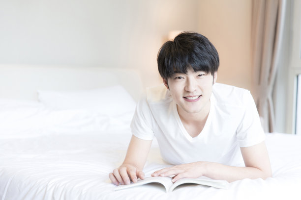 年轻的东亚男子摊开在现代家庭卧室的床上阅读一本书-库存照片图片下载