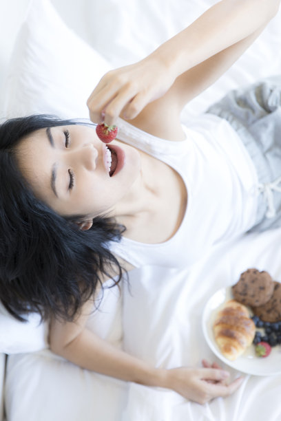 年轻的东亚妇女吃甜点，坐在床上的现代家庭卧室-库存照片图片下载