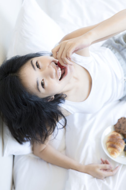 年轻的东亚妇女吃甜点，坐在床上的现代家庭卧室-库存照片图片下载