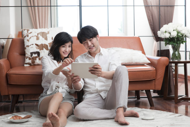 年轻的东亚夫妇坐在客厅地毯上使用平板电脑-库存照片图片下载
