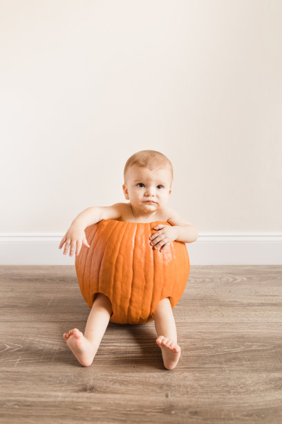2022年秋天，一个可爱的10个月大的男婴坐在一个真正的橙色南瓜里，腿和胳膊在家里的地板上图片下载