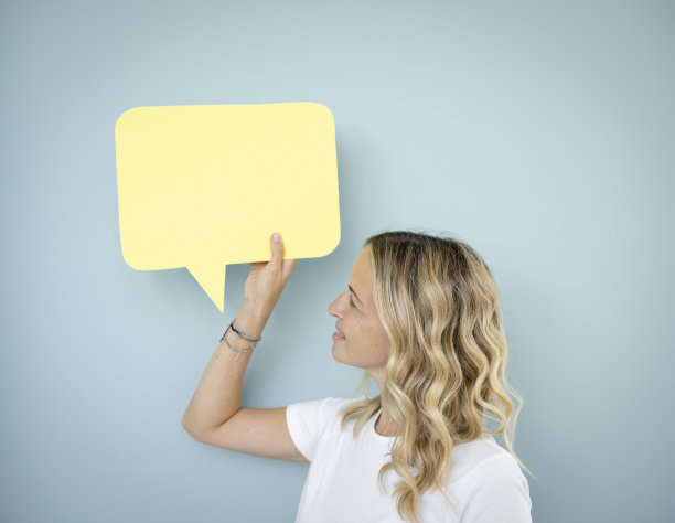 漂亮的金发卷发的年轻女子站在蓝色的墙前，拿着演讲泡沫图片下载