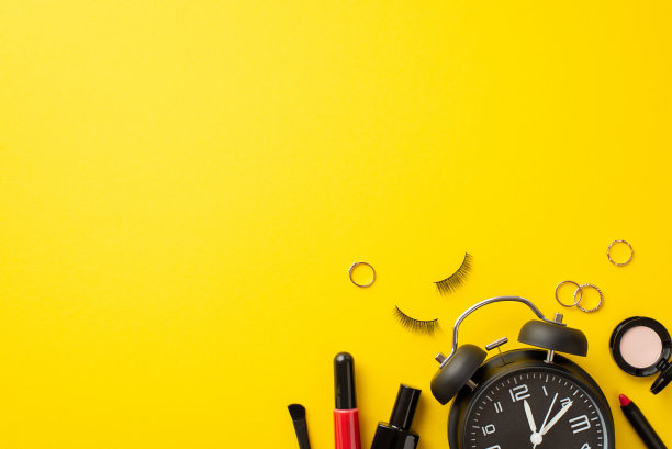 星期一的概念。闹钟化妆品的俯视图照片，指甲油，假睫毛，眼影，唇彩，刷子和金戒指在孤立的黄色背景与copyspace图片下载