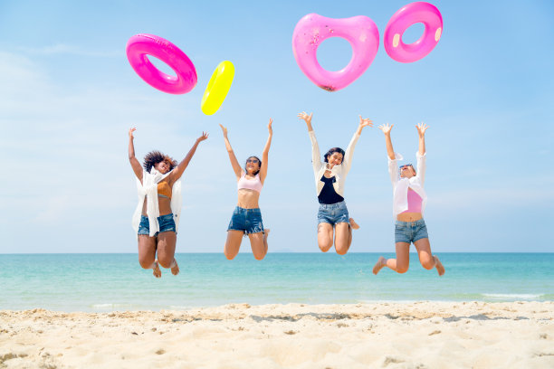四个十几岁的小女孩在蓝天碧水的沙滩上跳着抛着五颜六色的游泳圈，在假期里一起玩得很开心。图片下载