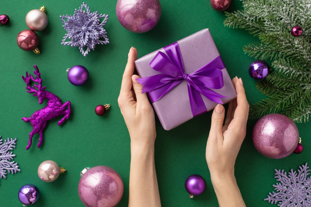 圣诞夜的概念。第一人称俯视图照片的女性手丁香礼品盒与紫罗兰蝴蝶结粉红色紫色小饰物驯鹿花雪花饰品杉树枝孤立的绿色背景图片下载