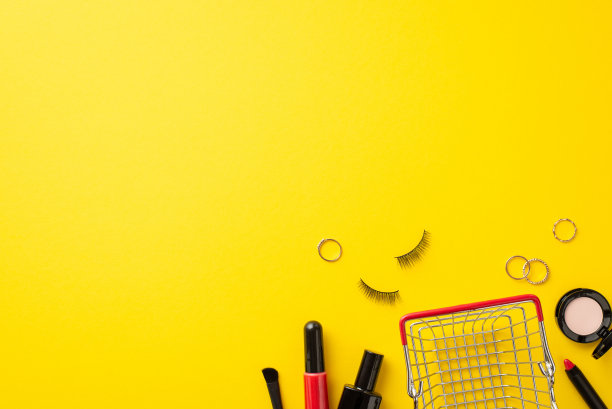 星期一的概念。购物车的俯视图照片，化妆品，指甲油，假睫毛，眼影，唇彩，刷子和金戒指在孤立的黄色背景与空白图片下载