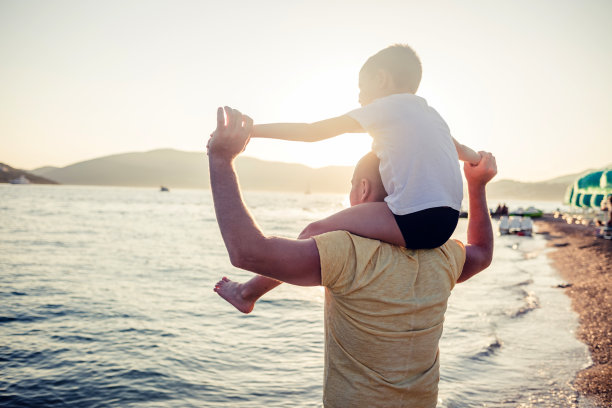 可爱的白人小男孩从后面欢呼，张开双臂坐在他父亲的肩膀上，在海滩上。图片下载