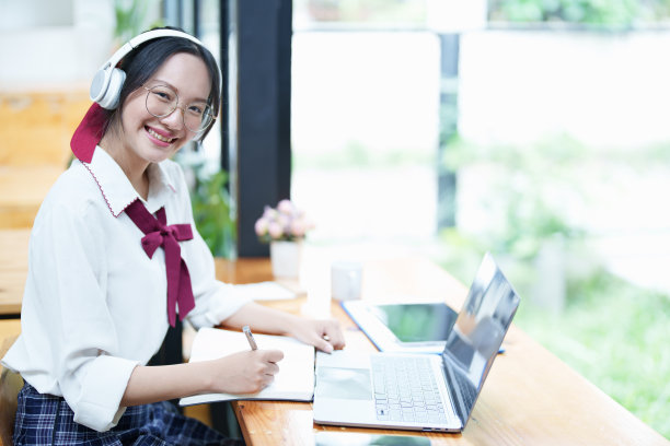 一名亚洲少女在图书馆的木桌上使用电脑，戴着耳机，用笔记本通过视频会议在线学习图片下载