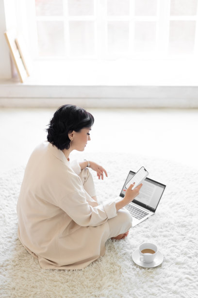 一个中年妇女坐在柔软的地毯上，手里拿着一个小玩意儿。对社交网络的依赖。为一家笔记本电脑公司远程工作。副本的空间。图片下载