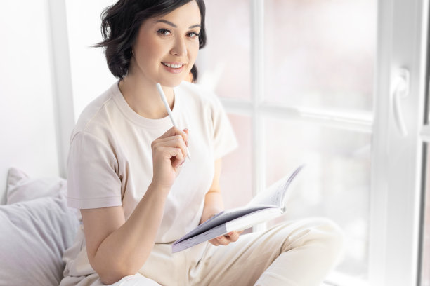 快乐微笑的褐发老太太在她的笔记本上做笔记。手里拿着一支白色的钢笔。坐在明亮的室内靠窗。看着相机。图片下载