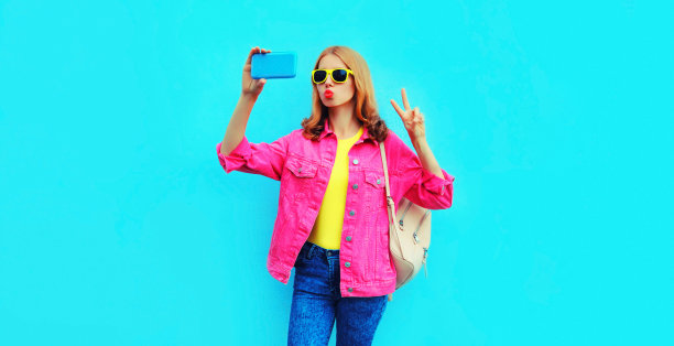 肖像快乐微笑时尚的年轻女子用智能手机自拍，吹她的嘴唇送吻，穿着粉红色的夹克，背包，蓝色背景的黄色太阳镜图片下载