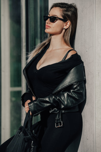 时尚的年轻女子长发，穿着结实的长袖紧身连体衣，黑色皮夹克和太阳镜摆姿势图片下载