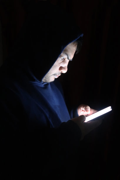 男性的形象，亚洲电话黑客穿帽衫在黑暗，脸照亮的智能手机屏幕，个人资料查看，互联网犯罪的概念图片下载