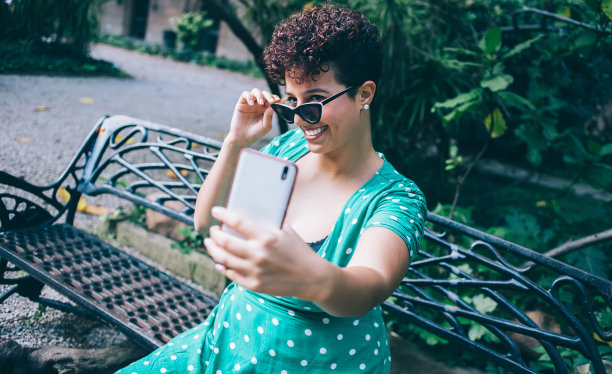 一位优雅的女士在公园的长椅上摸着墨镜，用智能手机自拍图片下载