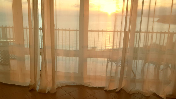 4k视频，金色的日落时分，透过阳台大全景窗上的窗帘，美丽的景色，大海在背景中图片下载