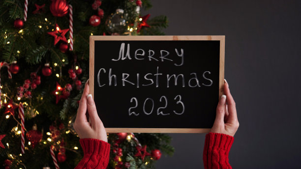 结婚圣诞横幅的概念。穿着红色毛衣的女性手拿黑板，上面写着2023年圣诞快乐，背景是用红色玩具和花环装饰的美丽的节日圣诞树图片下载