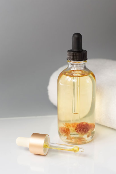 在灰色背景的白色毛巾布旁边，放着一瓶有机化妆品油，上面有花蕾和一个分配器。图片下载