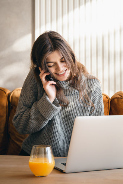 迷人的年轻女子一边打电话一边用笔记本电脑工作。图片下载