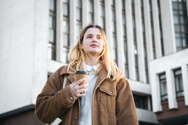 在大学的背景下，一个时髦的年轻女子端着一杯咖啡。图片下载