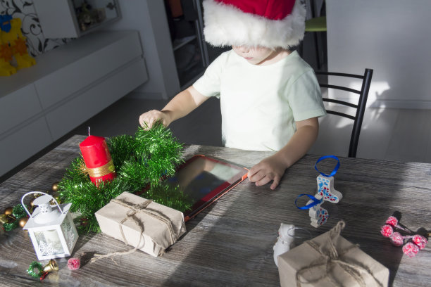 一个小孩子戴着圣诞老人的帽子，手里拿着圣诞礼物，坐在新年的桌子旁，手里拿着平板电脑。图片下载
